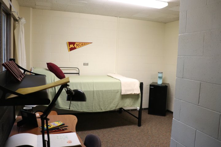 A college dorm room with a bed against the wall, neatly made with green bedding, a small fridge with a water bottle on top, and a desk with drawing supplies. A University of Minnesota Morris pennant hangs on the wall.